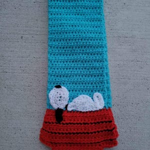 May include: A crocheted scarf with a red and black dog house design and a white crocheted dog on top. The scarf is turquoise blue.