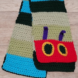 May include: A crocheted scarf with a colorful striped pattern in shades of green, blue, and beige. A red crocheted appliqué of a tractor with purple exhaust pipes and yellow wheels is attached to the scarf.
