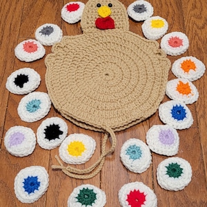 May include: A tan crochet chicken-shaped hot pad with a red comb and yellow beak, surrounded by crochet egg-shaped coasters in various colors. The hot pad has a loop for hanging.