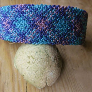 Puede incluir: Un brazalete tejido con un patrón de color azul, morado y rosa. El brazalete está hecho con una técnica de anudado y está sobre una superficie de madera marrón claro.