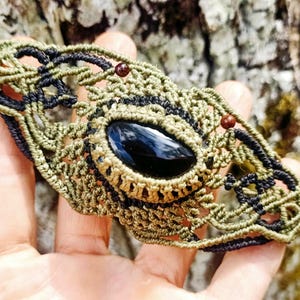 Puede incluir: Un brazalete de macramé verde y negro con una piedra preciosa negra y cuentas marrones.