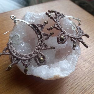 May include: A pair of brown macrame earrings with silver hoops and gold beads. The earrings are displayed on a white crystal.