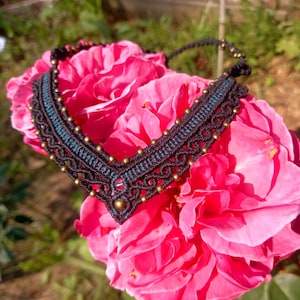 May include: A handcrafted black macrame necklace with a teal center and gold-colored beads. The necklace is displayed on a vibrant pink rose, creating a striking contrast. The intricate knotting and beadwork highlight the artisan craftsmanship.