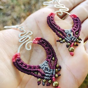 Peut inclure: Une paire de boucles d'oreilles faites à la main avec un anneau en fil d'argent et un motif en macramé dans des tons de violet, rose et gris. Les boucles d'oreilles sont ornées de petites perles dorées.