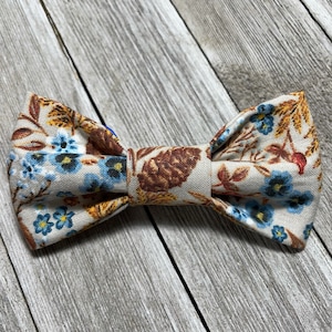 May include: A beige bow tie with a brown, blue, and yellow floral pattern. The bow tie is made of fabric and has a simple design.
