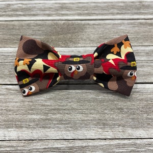 May include: A brown bow tie with a pattern of turkeys wearing pilgrim hats. The turkeys are brown, red, and white.