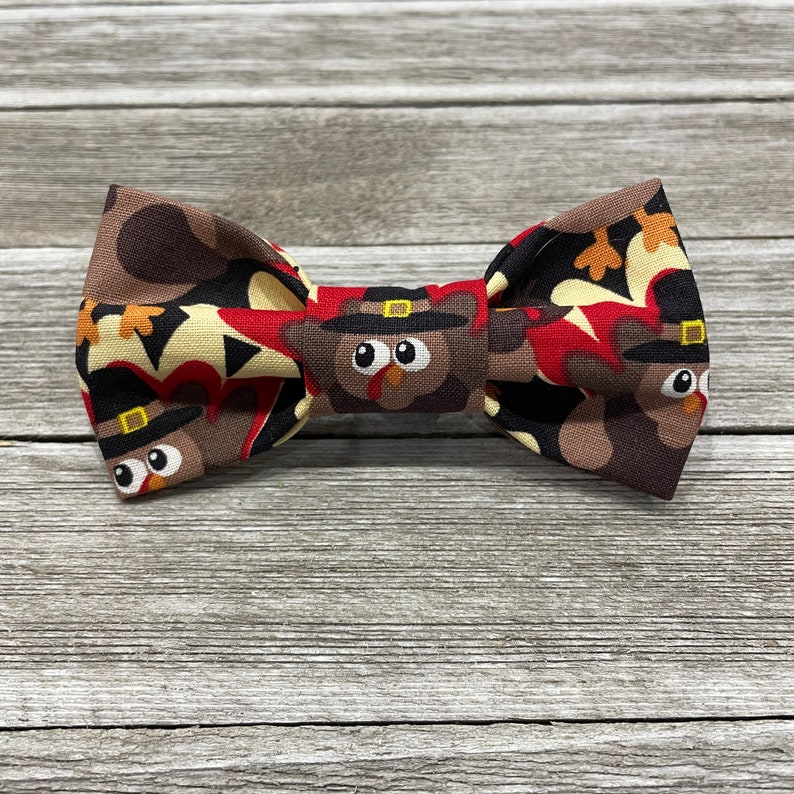 May include: A brown bow tie with a pattern of turkeys wearing pilgrim hats. The turkeys are brown, red, and white.
