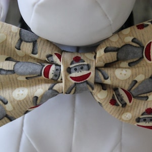May include: A tan bow tie with a pattern of gray sock monkeys wearing red hats. The monkeys are facing different directions and some are holding a white button.