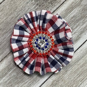 May include: A red, white, and blue plaid fabric flower with a large, sparkly rhinestone center.
