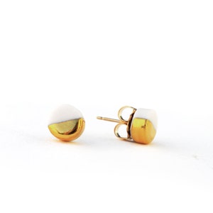 May include: A pair of gold and white ceramic stud earrings. The earrings are round and have a half-circle of gold on the bottom.