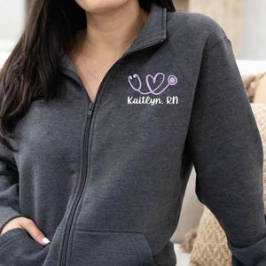 May include: A dark charcoal gray zip-up hoodie featuring a light purple stethoscope shaped into a heart on the left chest. Below the stethoscope graphic, the text "Kaitlyn. RN" is printed in white. The hoodie has a high collar and front pockets.