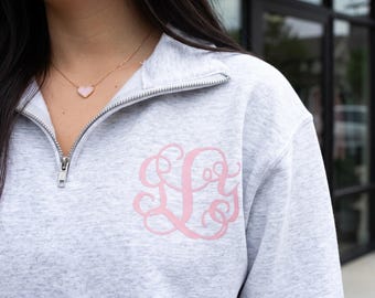 May include: A light gray heathered quarter-zip sweatshirt featuring a pink monogram design on the left chest. The garment has a silver-toned metal zipper at the collar and long sleeves. A delicate gold-toned necklace with a small pink heart pendant is worn.