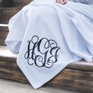 May include: A light gray throw blanket featuring a large, black, decorative script monogram with the letters H, G, and B intertwined in a circular design. The fabric has a soft, smooth texture and is draped over a set of wooden stairs.