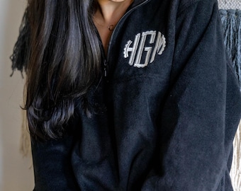 May include: A black fleece pullover featuring a quarter-zip neckline and a silver embroidered monogram on the left chest. The monogram consists of the letters HGM in a stylized font, surrounded by a circular scalloped border design.