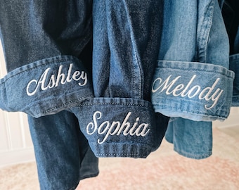 May include: Three denim shirts in varying shades of blue, each featuring a long-sleeved cuff embroidered with a name in white script. The cuffs display the names Ashley, Sophia, and Melody, showcasing different denim washes from dark to light.