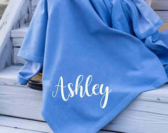 May include: A solid blue blanket featuring the name Ashley printed in white script lettering. The fabric is draped over the lap, showing a smooth texture and consistent color throughout the material, with the text clearly visible on the front.