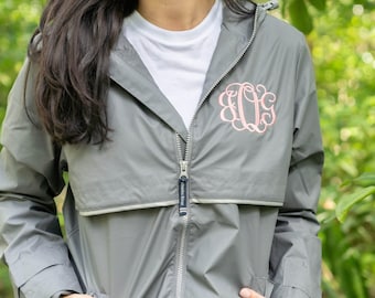 May include: A gray windbreaker jacket featuring a white undershirt and a pink embroidered monogram on the left chest. The jacket has a full-length front zipper with a dark pull tab and a horizontal seam detail across the upper chest area.