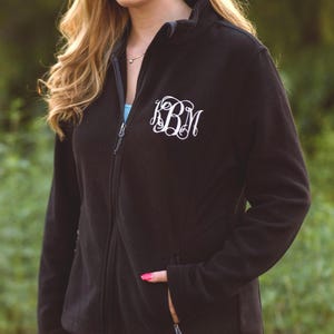 May include: A black fleece zip-up jacket featuring a white embroidered monogram with the letters KBM in a decorative script font on the upper left chest. The jacket has a high collar, long sleeves, and two side pockets with zipper closures.