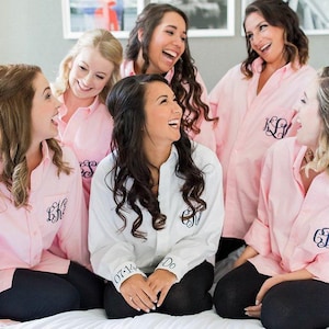 May include: A group of long-sleeved button-down shirts in light pink and white. The shirts feature dark-colored embroidered monograms on the chest. One white shirt displays the text 07-14 and I Do embroidered on the sleeve cuffs in dark thread.