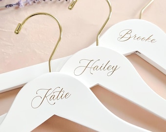 Puede incluir: Tres perchas de madera blanca con ganchos de metal dorado. Cada percha está personalizada con un nombre escrito en letra cursiva marrón: Katie, Hailey y Brooke. Las perchas están dispuestas en una formación escalonada y superpuesta.