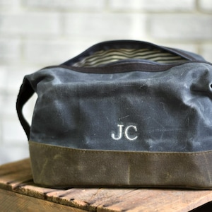 May include: A charcoal gray waxed canvas toiletry bag featuring a dark brown base and the embroidered initials JC in white thread. The bag has a wide zippered opening that reveals a black and white striped interior lining and side handles.