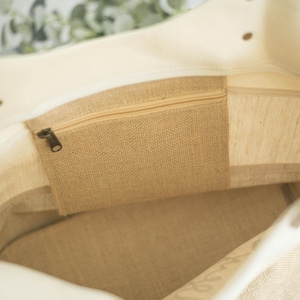 May include: Inside view of a beige tote bag with a zippered pocket made of burlap. The bag has a light-colored canvas exterior and brown leather handles. The interior features a burlap pocket and a canvas lining.