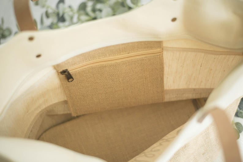 May include: Inside view of a beige tote bag with a zippered pocket made of burlap. The bag has a light-colored canvas exterior and brown leather handles. The interior features a burlap pocket and a canvas lining.