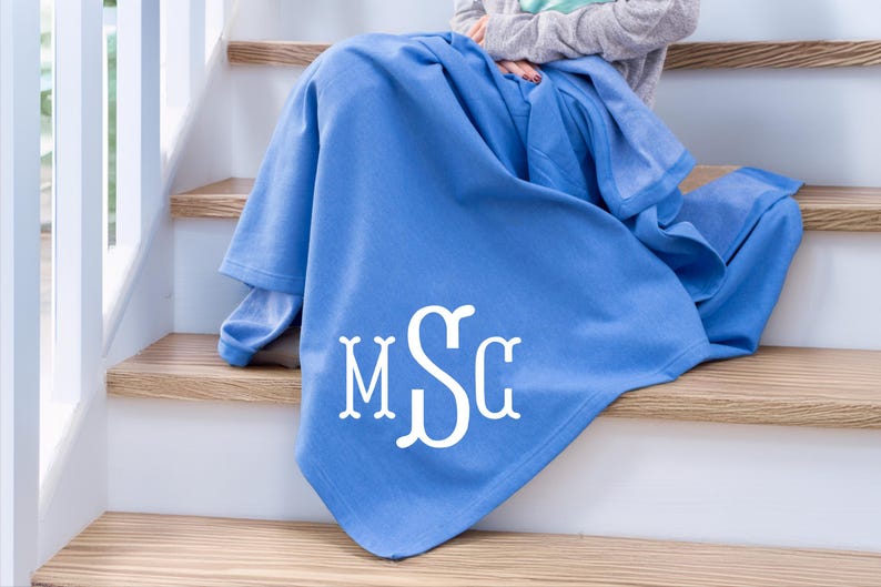 May include: A solid cornflower blue throw blanket featuring a large, white, decorative three-letter monogram with the letters M, S, and G in a stylized serif font centered on the lower portion of the fabric, draped over a set of wooden stairs.