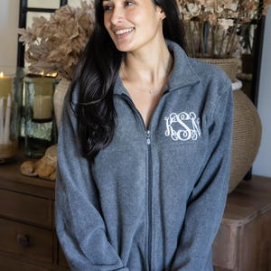 May include: A charcoal gray fleece jacket with a full-length front zipper and a high collar. The left chest features an embroidered white monogram with the letters KSN in a decorative, swirling script font.