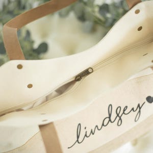 May include: Cream-colored tote bag with brown leather handles and accents. The bag features a zipper closure and the name "lindsey" in a cursive font with a heart symbol. Gold rivets secure the handles.
