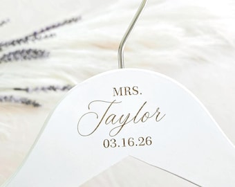 May include: A white wooden clothes hanger with a silver metal hook. The hanger features the engraved text MRS. Taylor and the date 03.16.26 in a dark brown script and serif font centered on the front of the curved wooden frame.