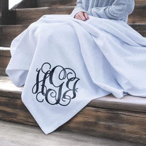 May include: A light gray throw blanket featuring a large, black, decorative script monogram with the letters H, G, and B intertwined in a circular design. The fabric has a soft, smooth texture and is draped over a set of wooden stairs.