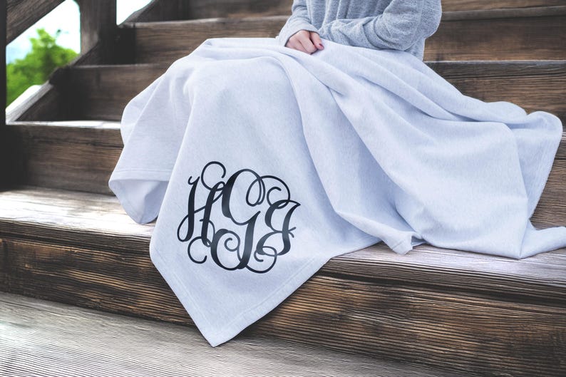 May include: A light gray throw blanket featuring a large, black, decorative script monogram with the letters H, G, and B intertwined in a circular design. The fabric has a soft, smooth texture and is draped over a set of wooden stairs.