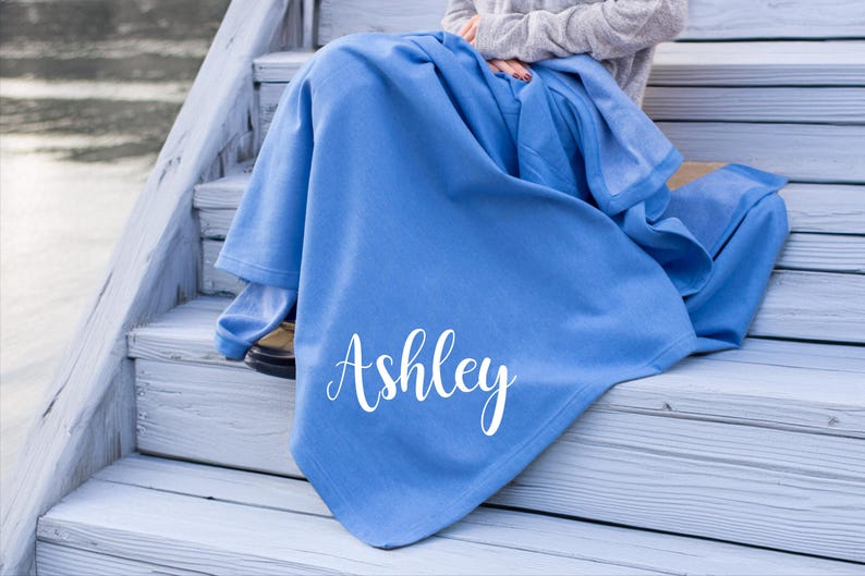 May include: A solid cornflower blue blanket featuring the name Ashley printed in a white cursive script font. The fabric appears soft and smooth, draped over the lap, covering the lower body and legs while extending down to the surface below.