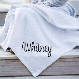 May include: A light gray throw blanket featuring the name Whitney printed in a black cursive script font. The fabric has a soft, smooth texture and is draped in folds, showcasing the personalized lettering clearly on the front surface.
