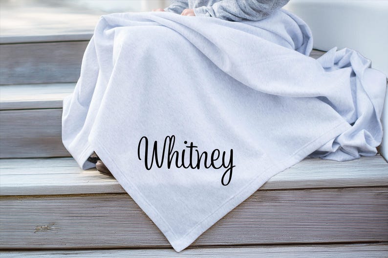 May include: A light gray throw blanket featuring the name Whitney printed in a black cursive script font. The fabric has a soft, smooth texture and is draped in folds, showcasing the personalized lettering clearly on the front surface.