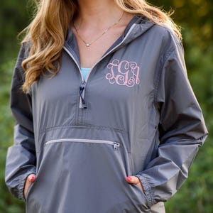 May include: A gray hooded pullover jacket featuring a quarter-zip neckline and a large front pocket with a zipper closure. The jacket is embroidered with a light pink monogram featuring the letters TGA in a decorative script font.