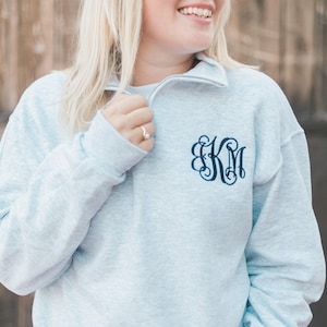 May include: A light blue heathered quarter-zip sweatshirt featuring a dark blue embroidered monogram on the left chest. The garment has long sleeves, a high collar with a zipper closure, and a relaxed fit with ribbed cuffs and hem.