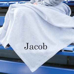 May include: A light gray blanket featuring the name Jacob printed in a black serif font. The blanket is draped over a surface, displaying a soft, folded texture across its surface. The fabric appears smooth and consistent in its light gray color.