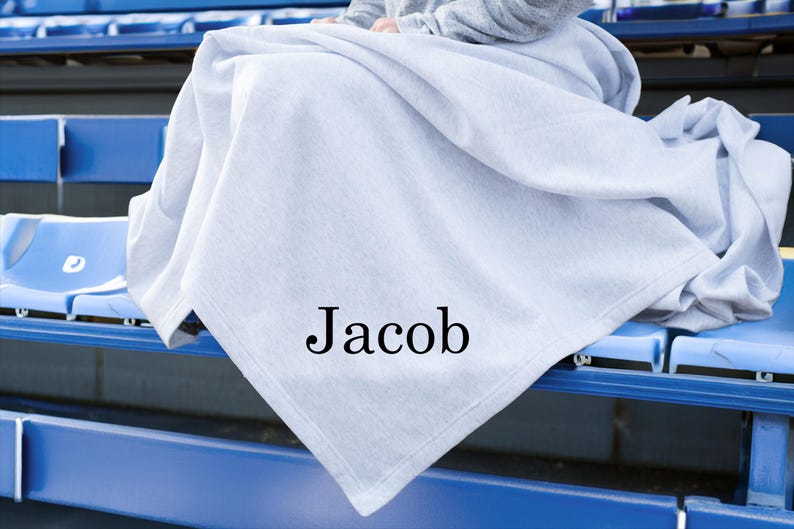 May include: A light gray blanket featuring the name Jacob printed in a black serif font. The blanket is draped over a surface, displaying a soft, folded texture across its surface. The fabric appears smooth and consistent in its light gray color.