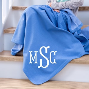 May include: A solid cornflower blue throw blanket featuring a large, white, decorative three-letter monogram with the letters M, S, and G in a stylized serif font centered on the lower portion of the fabric, draped over a set of wooden stairs.
