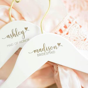 May include: Two white wooden clothes hangers with gold metal hooks. The hangers feature gold script text reading ashley MAID OF HONOR and madison BRIDESMAID, each accented with a small gold heart symbol above the names in a cursive font.