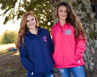 May include: A navy blue windbreaker with a pink monogram and a bright pink windbreaker with a teal monogram. Both jackets feature a front zipper, a hood, and side pockets, worn over blue denim jeans with distressed detailing.