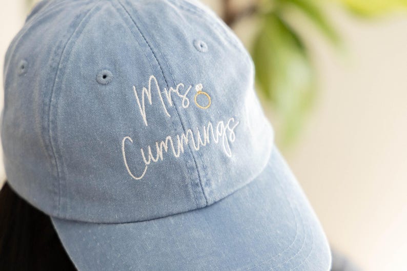 May include: A light blue baseball cap with white embroidered text that reads Mrs. Cummings, accompanied by a small gold-colored embroidered ring icon. The cap features a classic six-panel design with stitched eyelets and a curved brim.