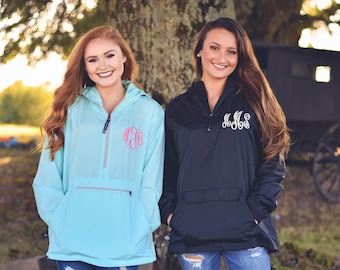 May include: A light blue pullover windbreaker with a pink monogram on the chest and a black pullover windbreaker with a white monogram on the chest. Both jackets feature a hood, a front pocket, and a quarter-zip closure at the neckline.