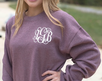 May include: A heathered mauve crewneck sweatshirt featuring a white, ornate, script-style monogram on the upper left chest. The garment has long sleeves, a ribbed neckline, and a soft, textured fabric finish throughout the design.