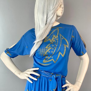 May include: A blue two-piece outfit with a gold abstract design. The top has short sleeves and a scoop neck. The skirt has a high waist and a tie belt.