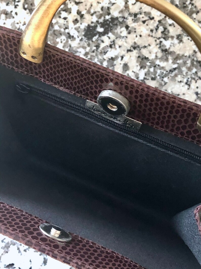 May include: A brown snakeskin handbag with a gold metal handle and a black fabric interior. The bag has a zipper closure and a metal clasp.