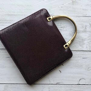 May include: A dark brown leather handbag with a gold-toned top handle. The bag has a textured, snakeskin-like pattern.