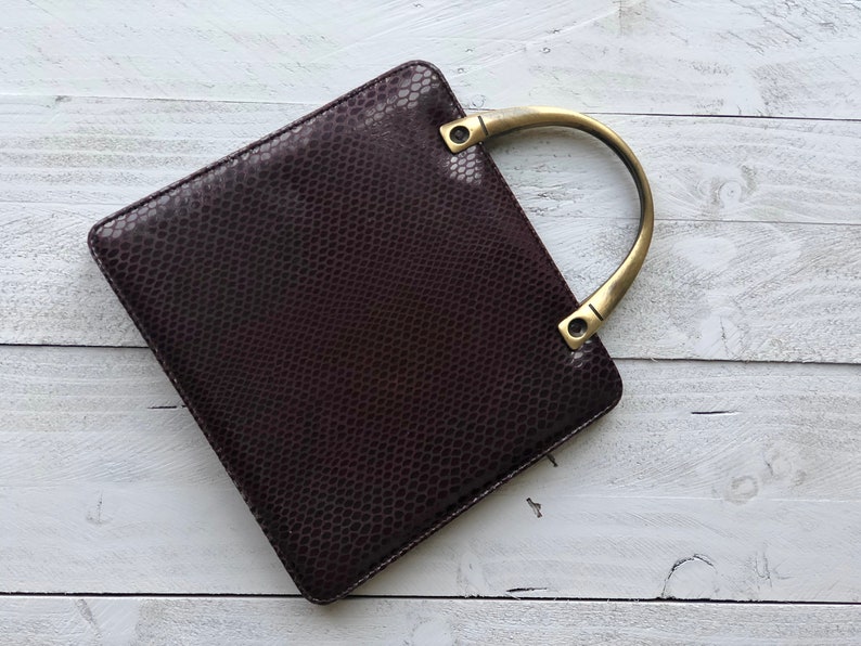 May include: A dark brown leather handbag with a gold-toned top handle. The bag has a textured, snakeskin-like pattern.